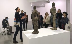 Un groupe de visiteurs et visiteuses regardent deux statues de l'exposition Hans Josephsohn, le conférencierdonne des information sur les oeuvres.